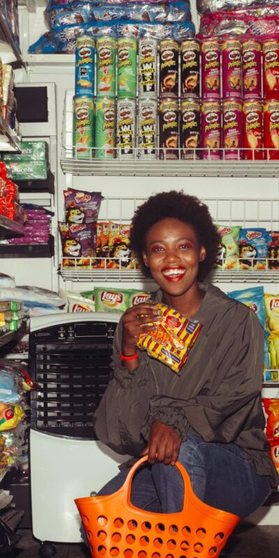 Happy black female in casual clothes with shopping bag demonstrating product squatting among grocery shelves while choosing goods in supermarket